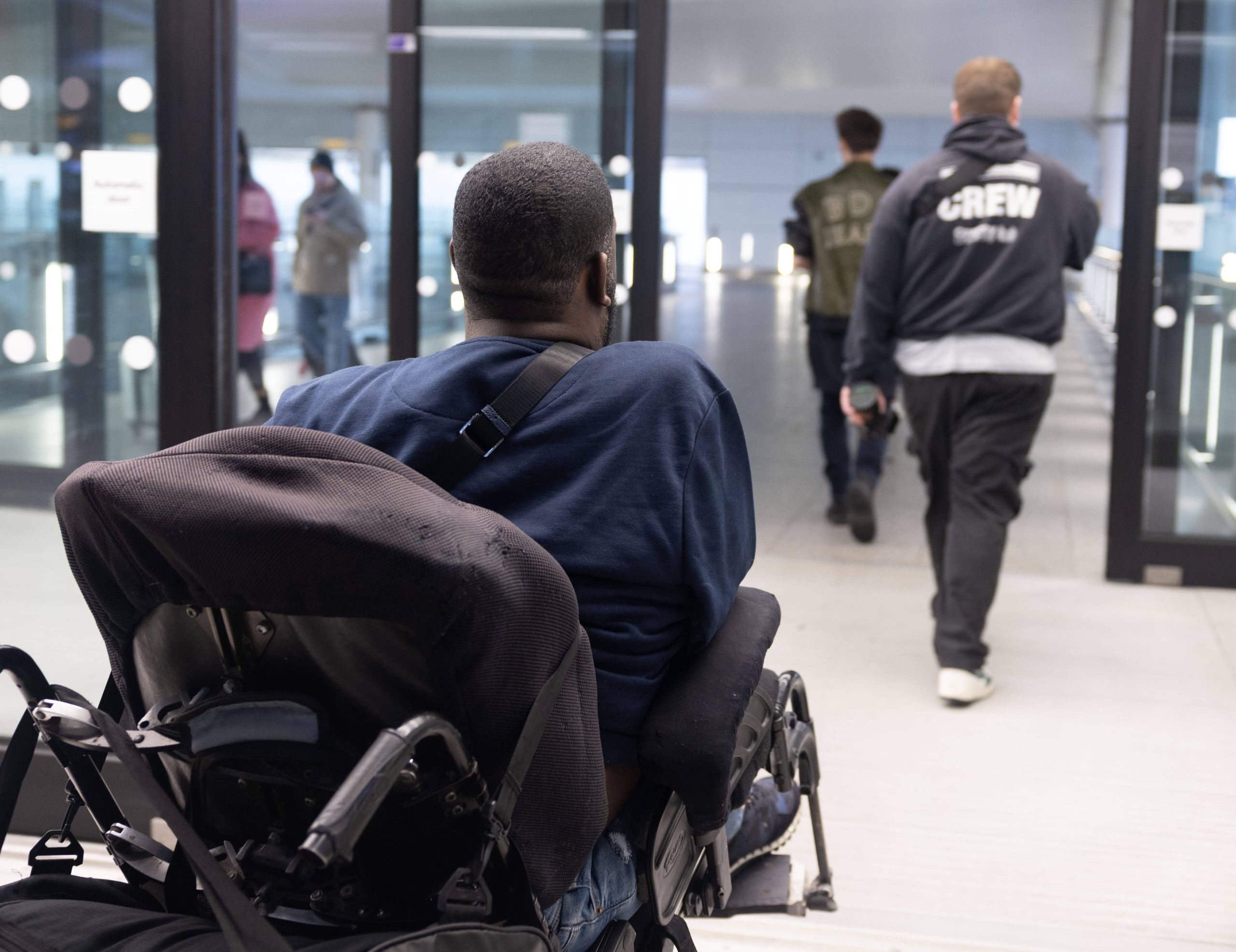 male wheelchair user going through automatic doors at Heathrow airport plus four people without mobility impairments on the concourse