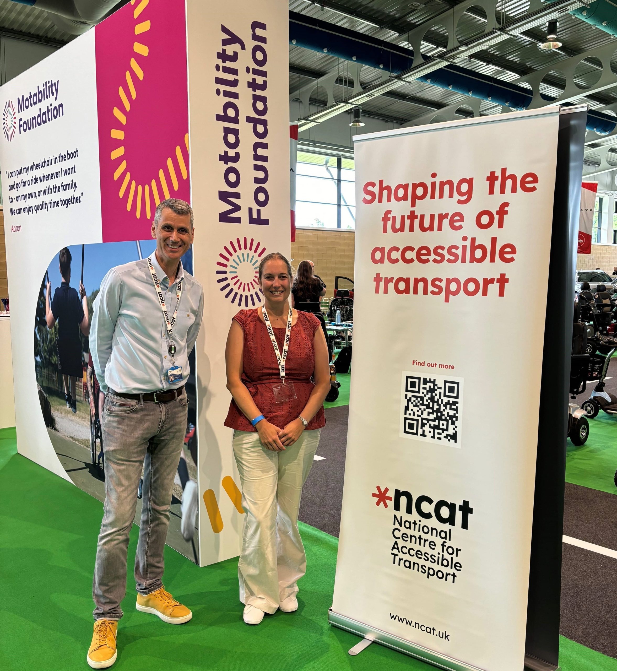 Paul Herriotts and Kay Atkin standing next to an ncat banner reading 'shaping the future of accessible transport'. A qr code is underneath the writing and above the ncat logo. This is in a large event space. Behind the banner there are some mobility vehicles and cars.