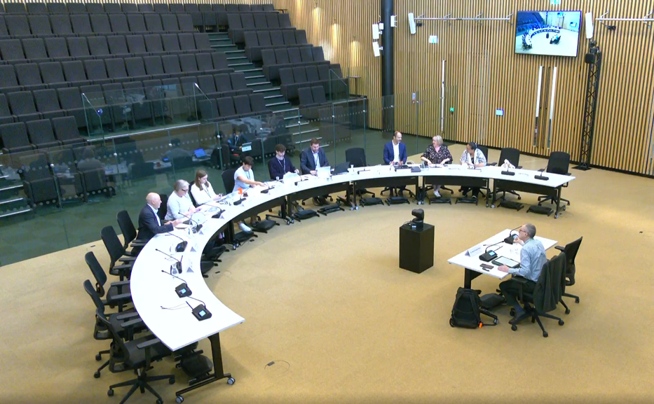 Tables arranged in a large semi circle with 18 chairs behind one side. Half the chairs are empty but nine are filled with a mix of men and women wearing office clothing. They are facing one small single table behind which one man is sitting. Behind all the tables are empty seats in lecture hall style.