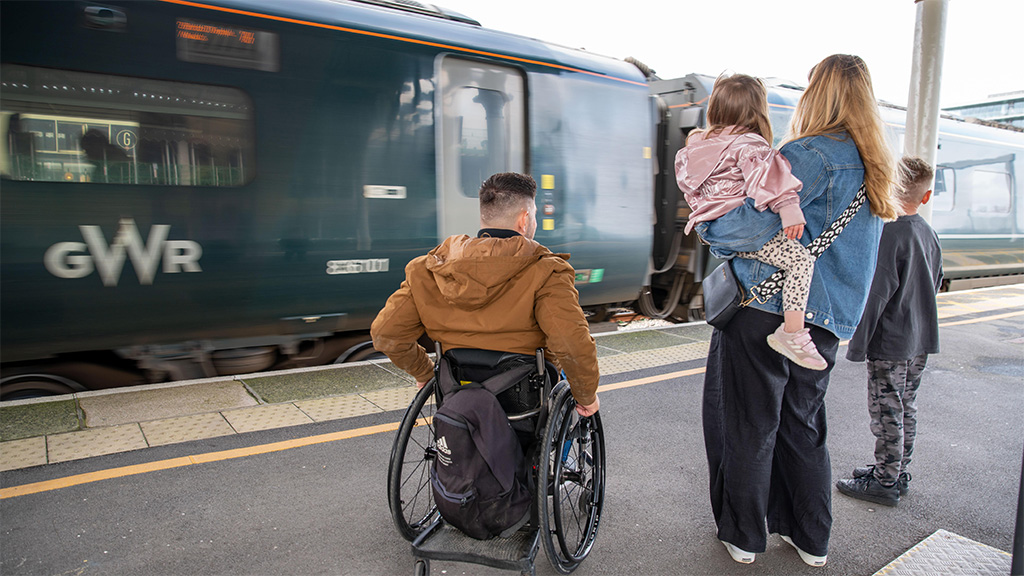 Family with their backs to the camera, waiting on a railway platform with train in front of them. One adult in a wheelchair, one adult holding a small cild, with another child alongside.
