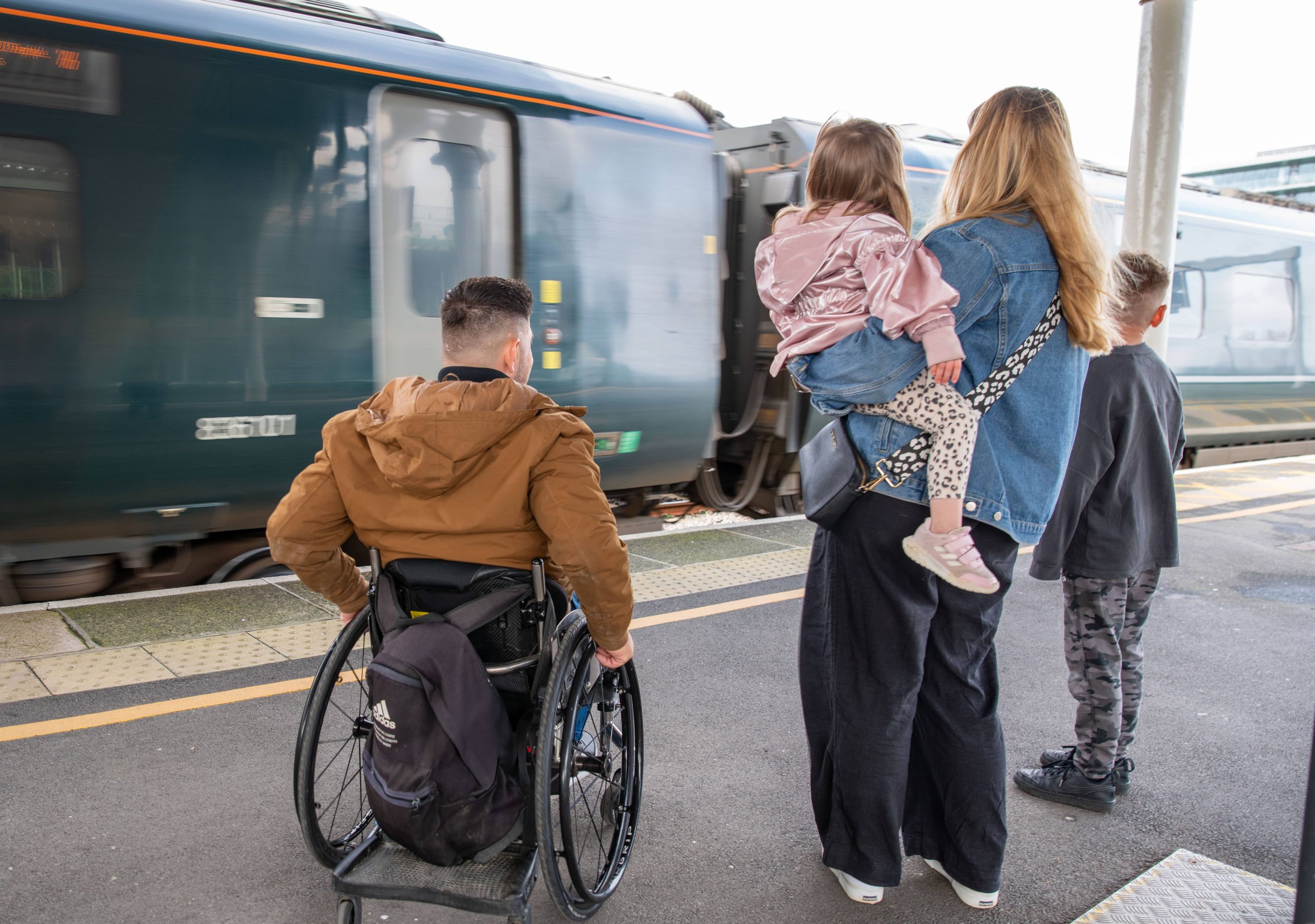 male wheelchair user and his partner and two children wait on train platform in from of a train