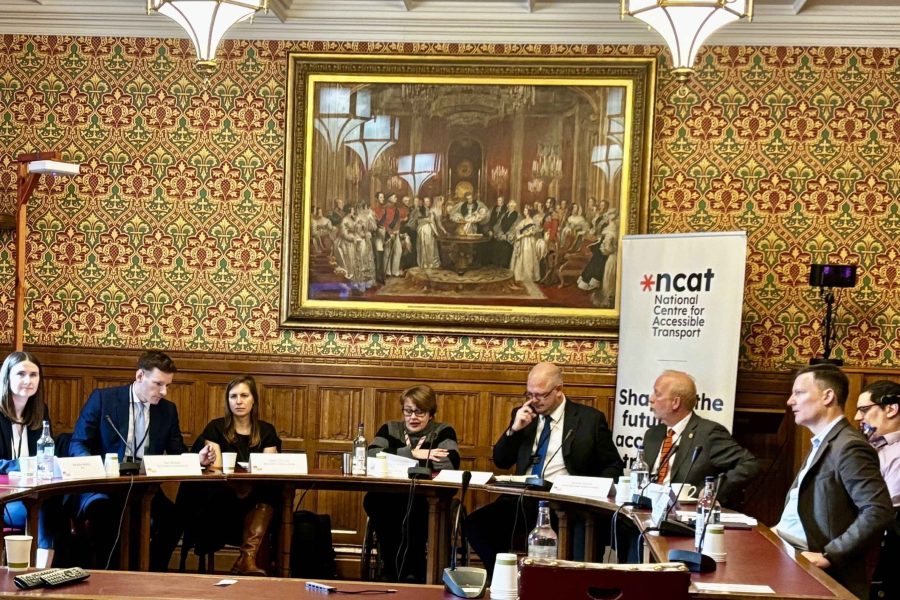 Baroness Tanni Grey-Thompson (centre) chairing Accessible Transport Policy Commission Event. There is a semi circle of tables with eight people (six male, two female) sitting behind them facing the camera. A standing poster sits behind them reading 'National Centre for Accessible Transport'. They are in the House of Lords and a very large old painting hangs on the wall behind them.