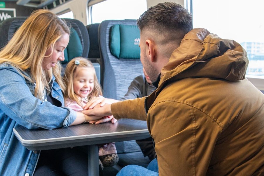 Male wheelchair user with his back to the camera faces his family (female partner and young children, one girl and one boy who is only visible by his arm.) They are sitting in a train carriage next to the window with a table between them.