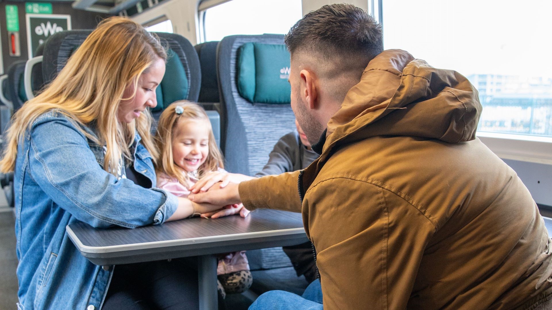 Male wheelchair user with his back to the camera faces his family (female partner and young children, one girl and one boy who is only visible by his arm.) They are sitting in a train carriage next to the window with a table between them.