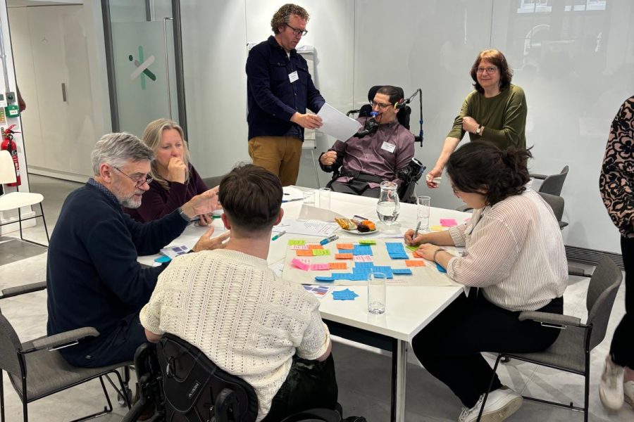 A group of people are sitting and standing round a table covered in sticky notes and pens in a plain white meeting room.